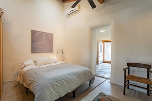 A bedroom with a bed, a chair, a wall-mounted air conditioner, and a door leading to another room. - Modern Historic 1900 Adobe Bungalow in Downtown - Low Summer Rates! 🌇 (Tucson)