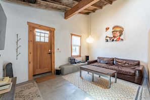 A cozy living room with a wooden door, a brown leather sofa, a wooden coffee table, and a patterned rug. - Modern Historic 1900 Adobe Bungalow in Downtown - Low Summer Rates! 🌇 (Tucson)