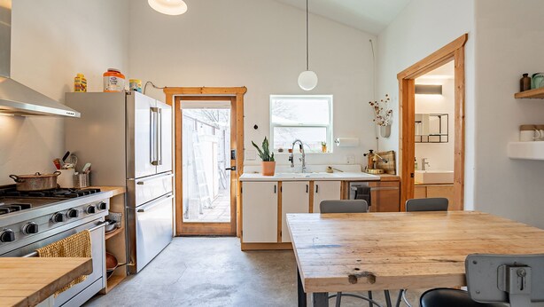 Una cocina moderna con una mesa de madera, electrodomésticos de acero inoxidable y vistas a través de una puerta abierta.