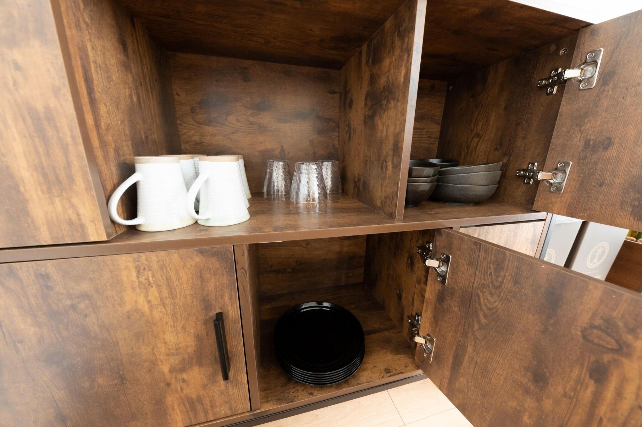 Un meuble en bois avec une étagère où sont posées des tasses blanches et des verres transparents, et un bol noir dans le compartiment inférieur.
