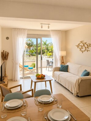 A modern living room with a sofa, dining table, and a view of a pool and palm trees. - Narcissos Nissi Beach apartment C2 (Ayia Napa)