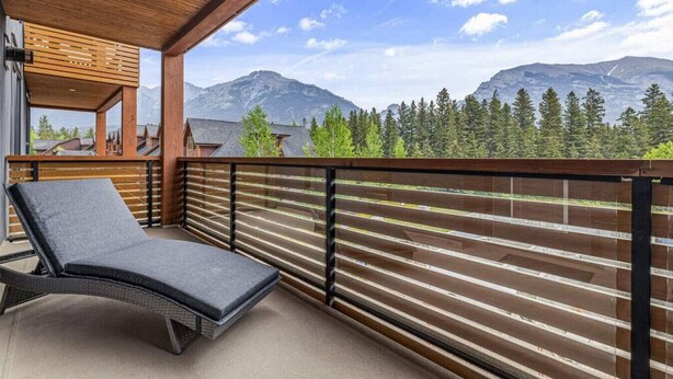 A balcony with a lounge chair, wooden railing, and a view of mountains and trees. - Stay in Style | Mountain Scene (Canmore)