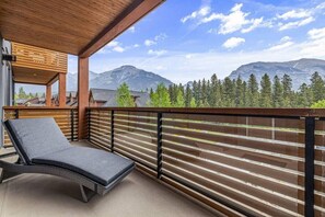 A balcony with a lounge chair, wooden railing, and a view of mountains and trees. - Stay in Style | Mountain Scene (Canmore)