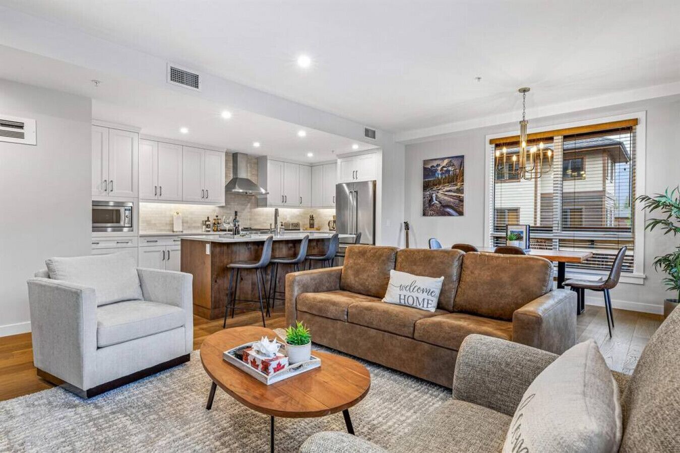 Una sala de estar moderna con un sofá, sillones y una mesa de centro. Se observa una zona de cocina con armarios blancos y una mesa de comedor con sillas.