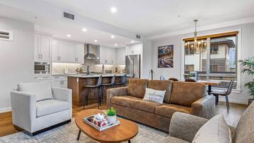 Una sala de estar moderna con un sofá, sillones y una mesa de centro. Se observa una zona de cocina con armarios blancos y una mesa de comedor con sillas.