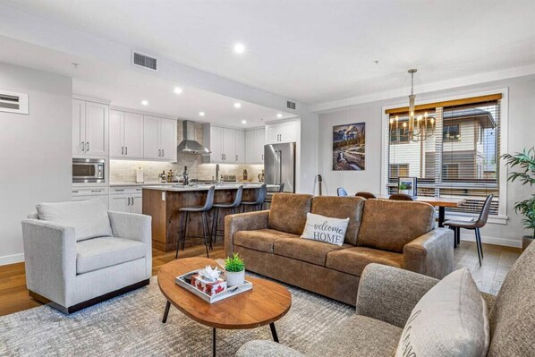 A modern living room with a sofa, armchairs, and a coffee table. There is a kitchen area with white cabinets and a dining table with chairs. - Stay in Style | Mountain Scene (Canmore)