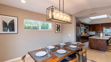A dining area with a wooden table set for six, a modern kitchen in the background, and a skylight above.