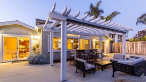 Een overdekt terras met buitenmeubilair, een pergola en uitzicht op het interieur van het huis.