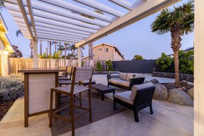 Un patio exterior con mobiliario de mimbre, una pérgola y vistas a una zona residencial.