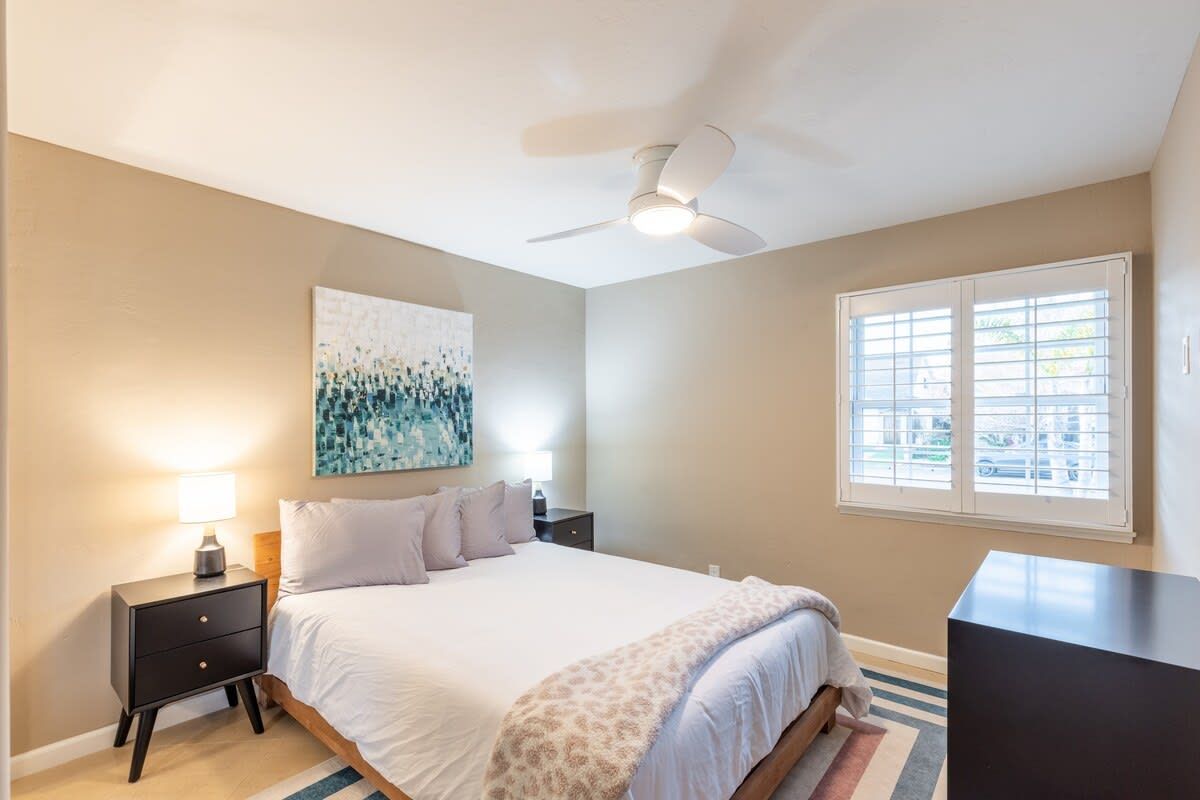 A bedroom with a bed, a nightstand, a desk, a ceiling fan, a window, and a painting on the wall.