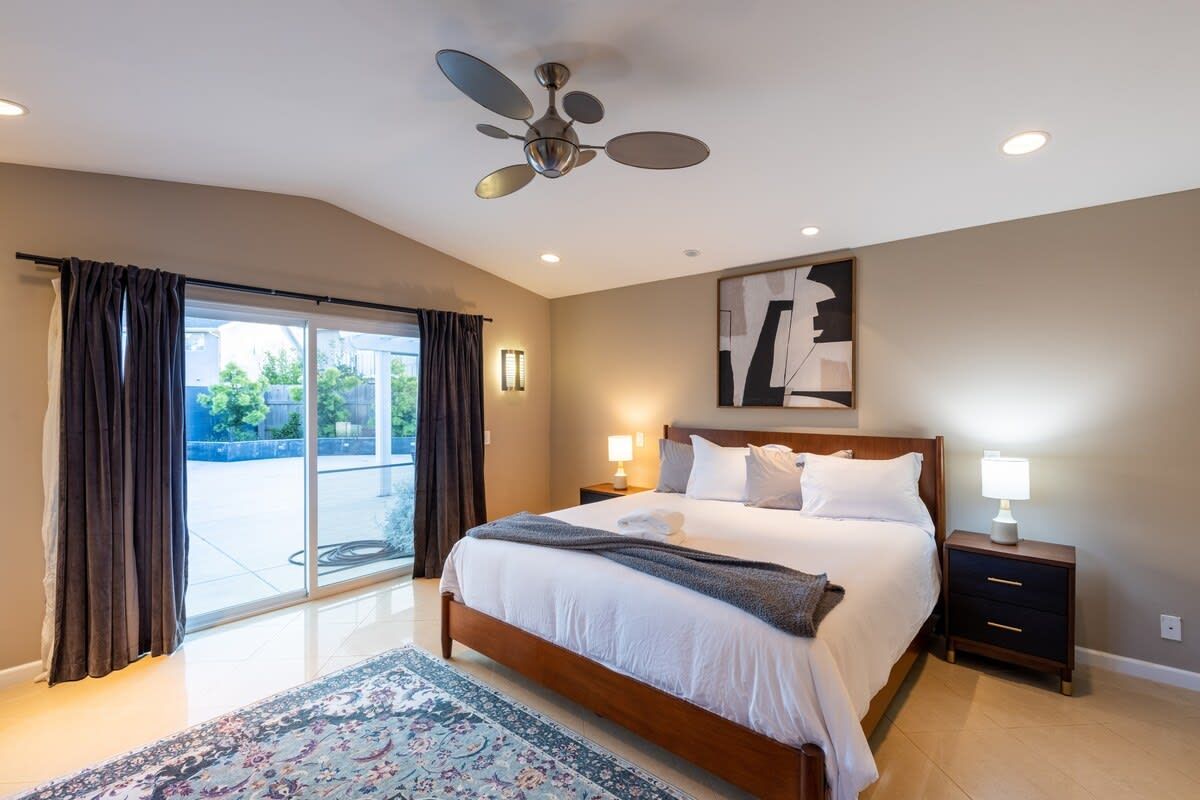 A hotel bedroom with a large bed, a ceiling fan, a view through the glass door, and a nightstand with a lamp.