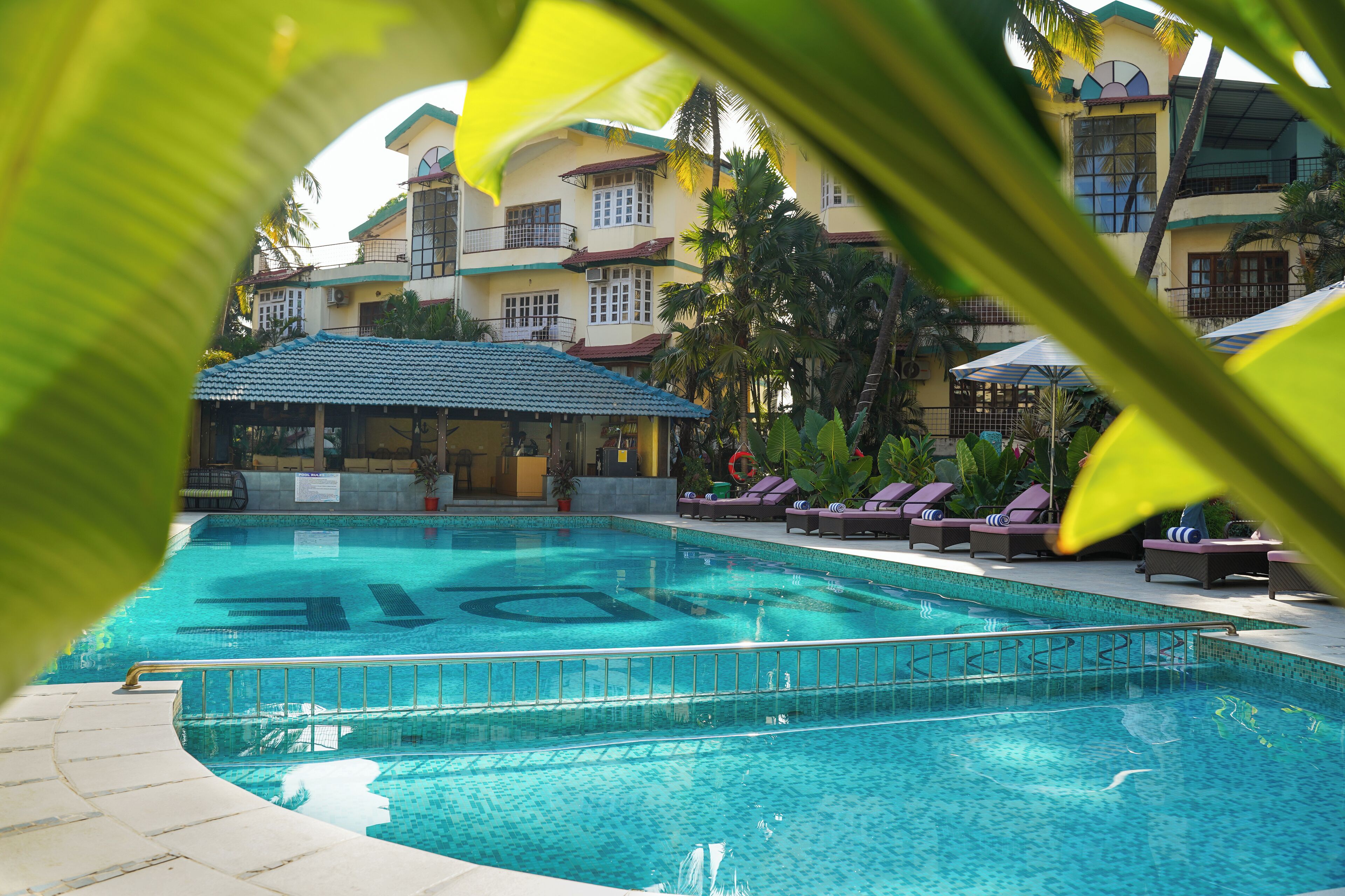 A swimming pool with lounge chairs and umbrellas, surrounded by a building with multiple balconies and palm trees.
