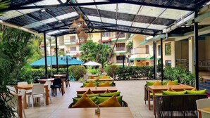 An outdoor patio with tables and chairs, covered by a metal roof, surrounded by greenery. - Indie Stays Goa (Candolim)