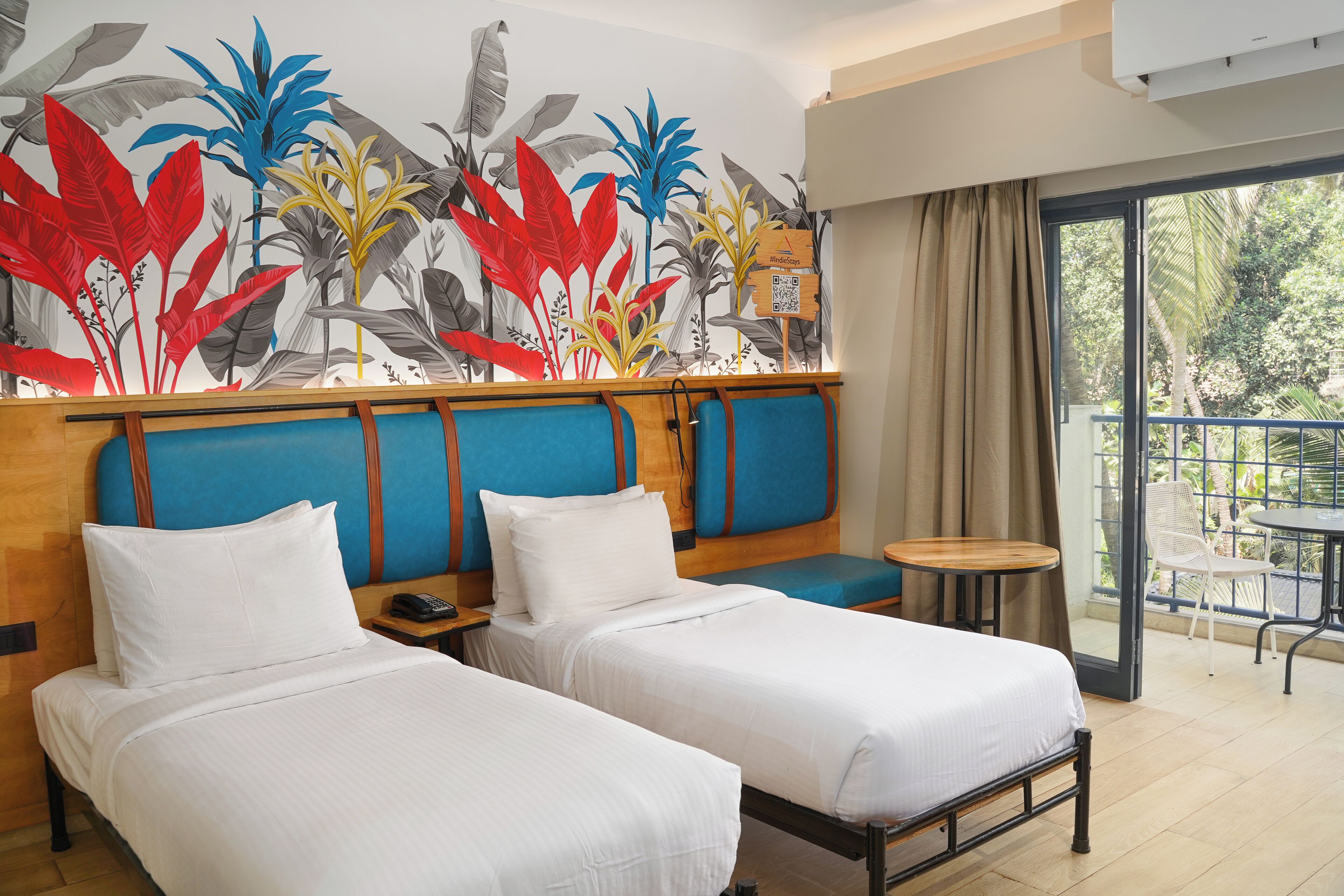 A hotel room with two beds, a mural of tropical plants, a balcony with a table and chairs, and a sliding glass door to the outside.