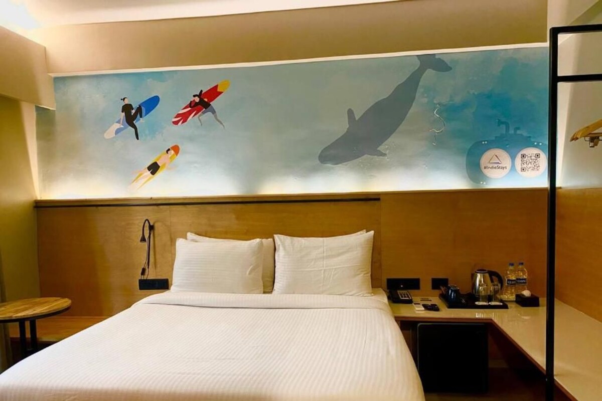 A hotel room with a large bed, a nightstand, a desk, and a wall mural depicting surfers and a whale.