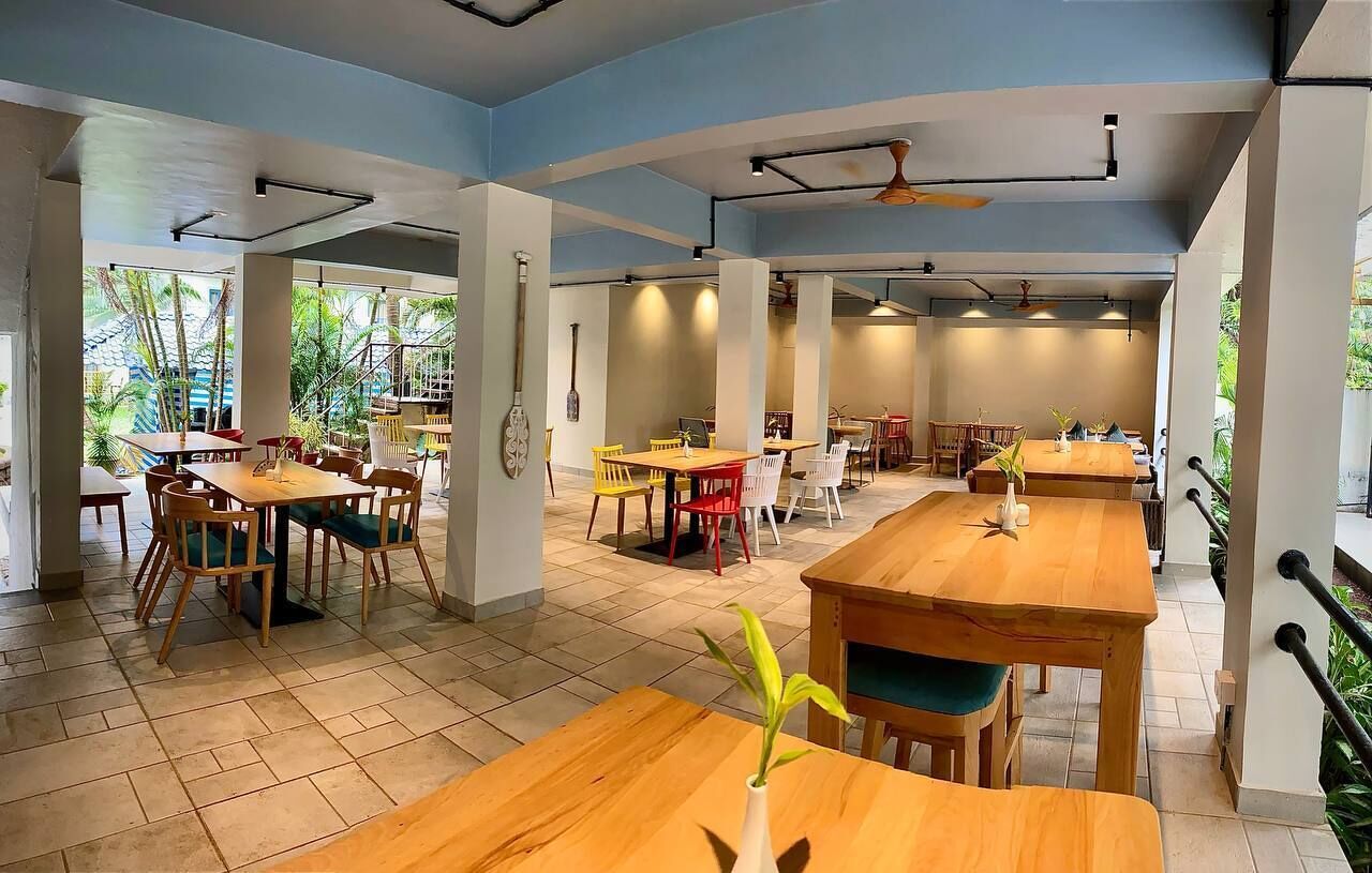 Un amplio comedor interior con varias mesas y sillas de madera, ventanas grandes que dejan entrar luz natural y un ventilador de techo.