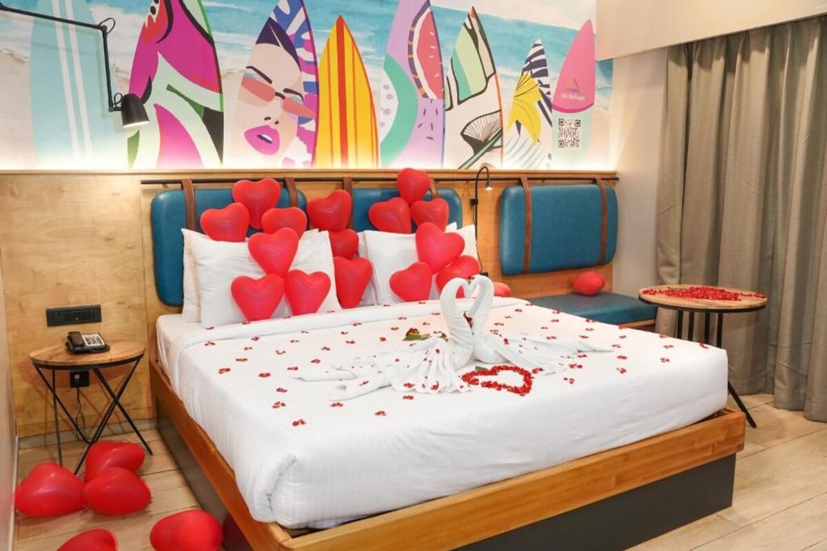 A hotel room with a bed adorned with red heart-shaped balloons, a swan-shaped pillow, and rose petals.