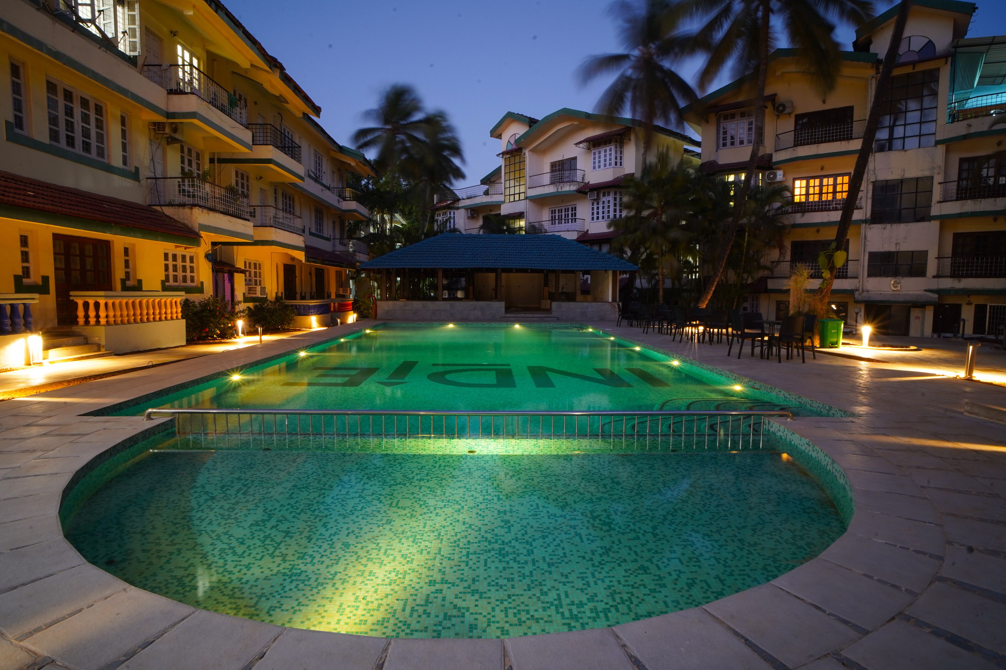 A swimming pool with a tiled deck, surrounded by multi-story buildings with balconies and palm trees.