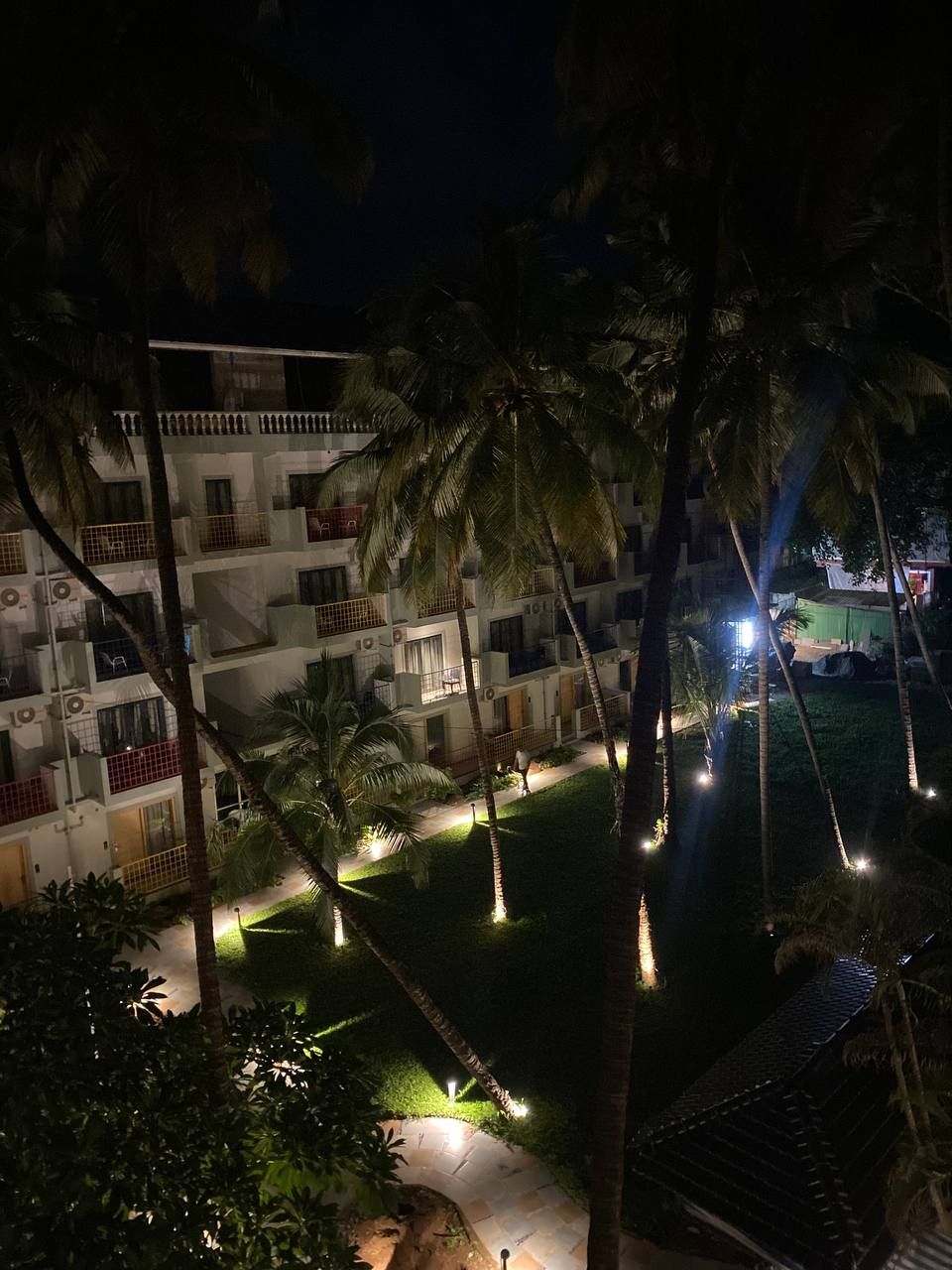 A multi-story building with balconies, surrounded by palm trees and illuminated pathways.
