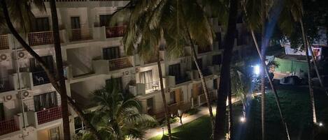 A multi-story building with balconies, surrounded by palm trees and illuminated pathways.