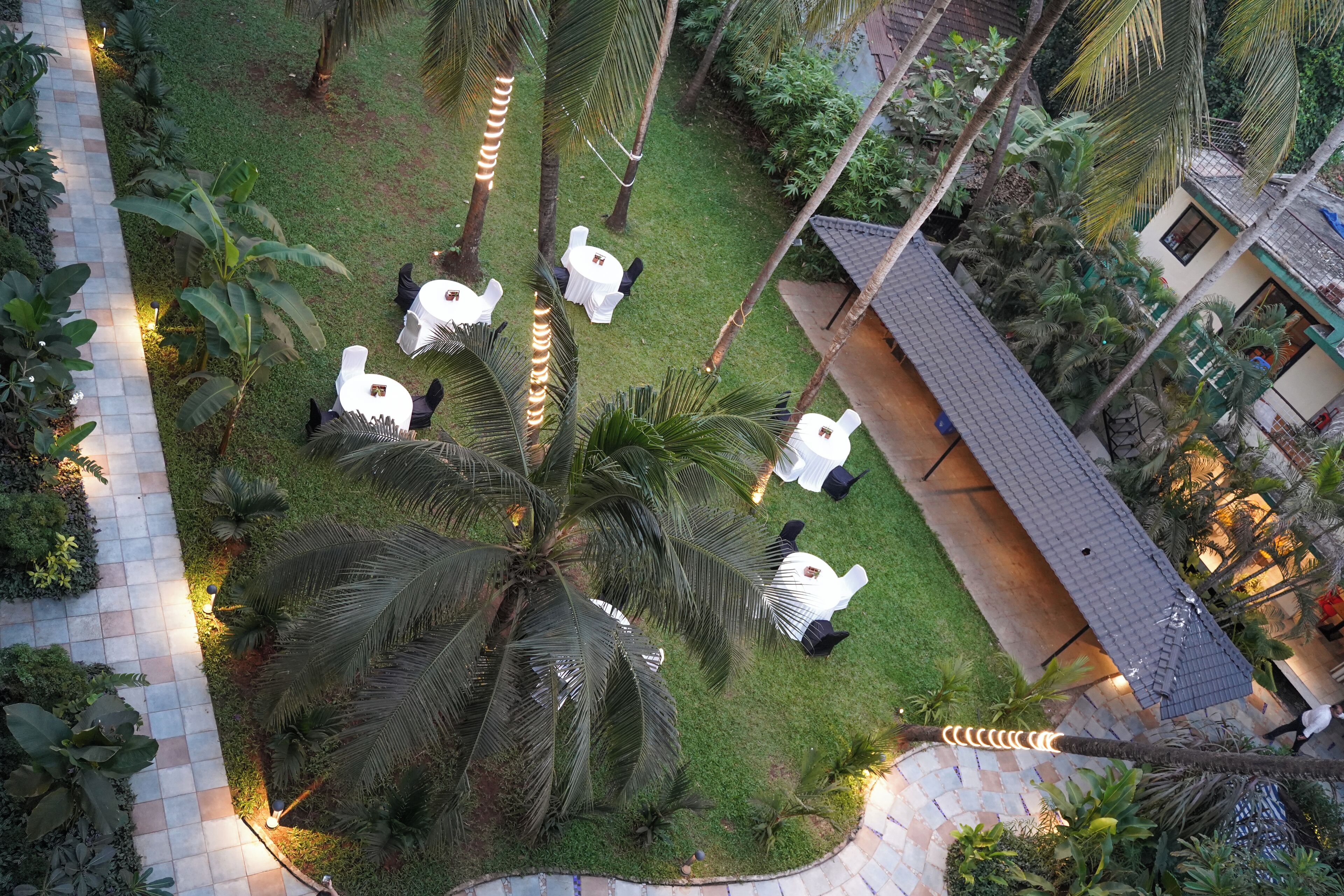 An aerial view of a garden with white tables and chairs arranged for outdoor dining, surrounded by lush greenery and palm trees.