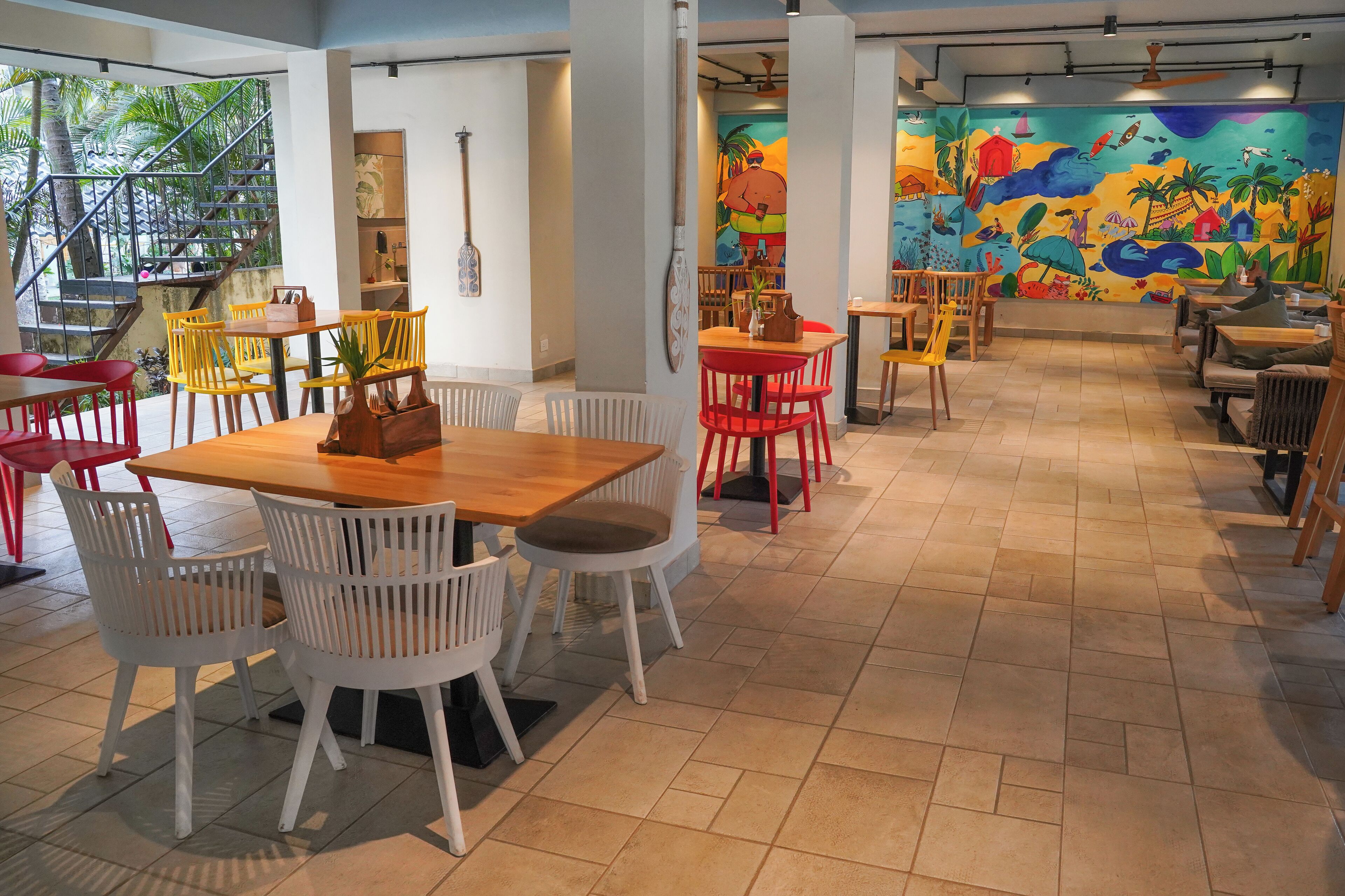 A restaurant with multiple dining areas, including a central table with white chairs and a wooden table with yellow chairs. There are red and white chairs around the tables, and a colorful mural on the wall.