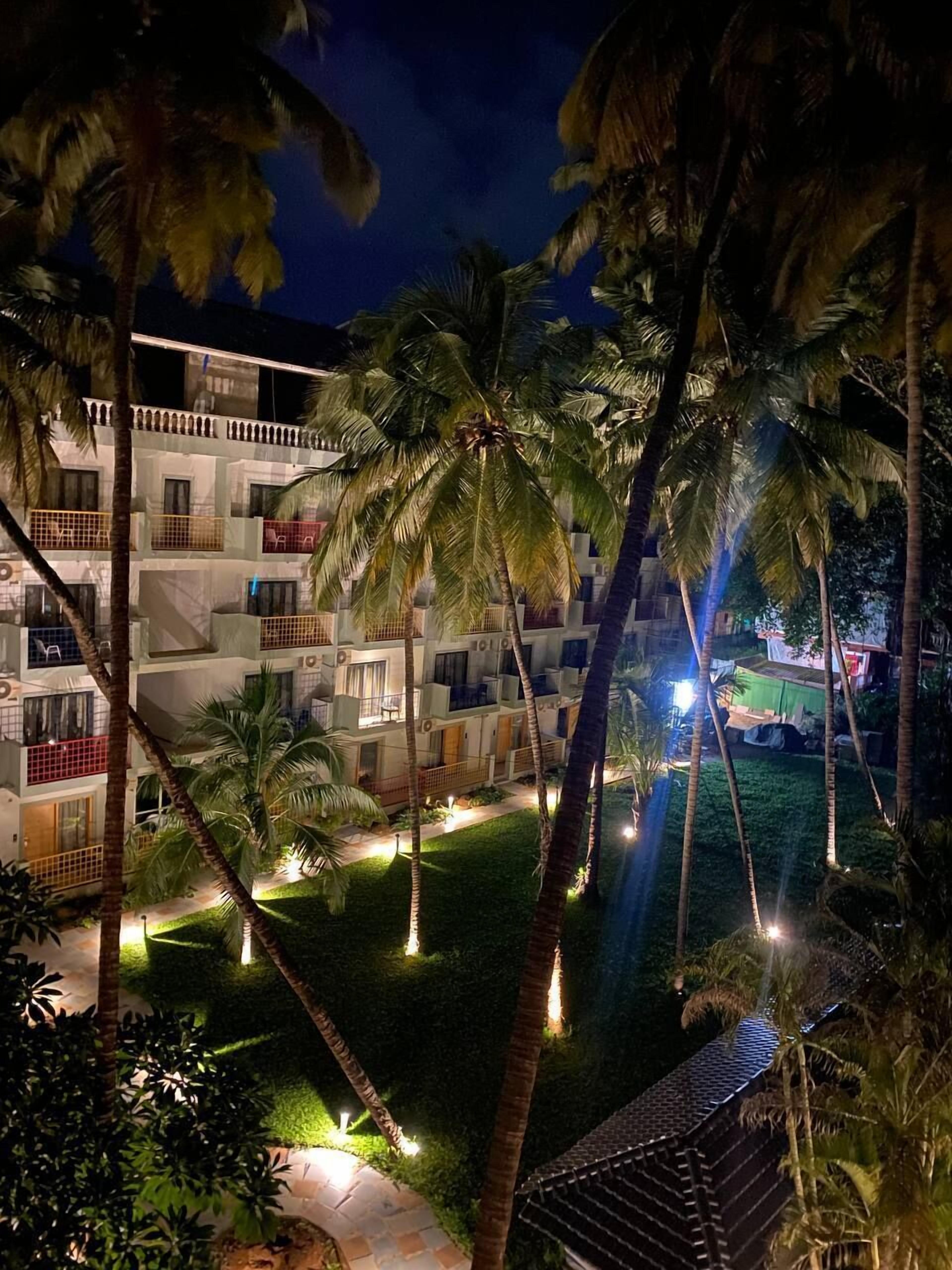 Un hotel de varios pisos con balcones, palmeras y un jardín bien iluminado.