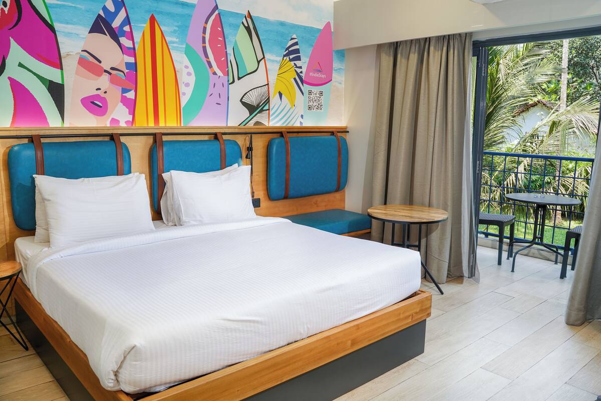 A hotel room with a large bed, a small table, and a balcony with a view of palm trees.