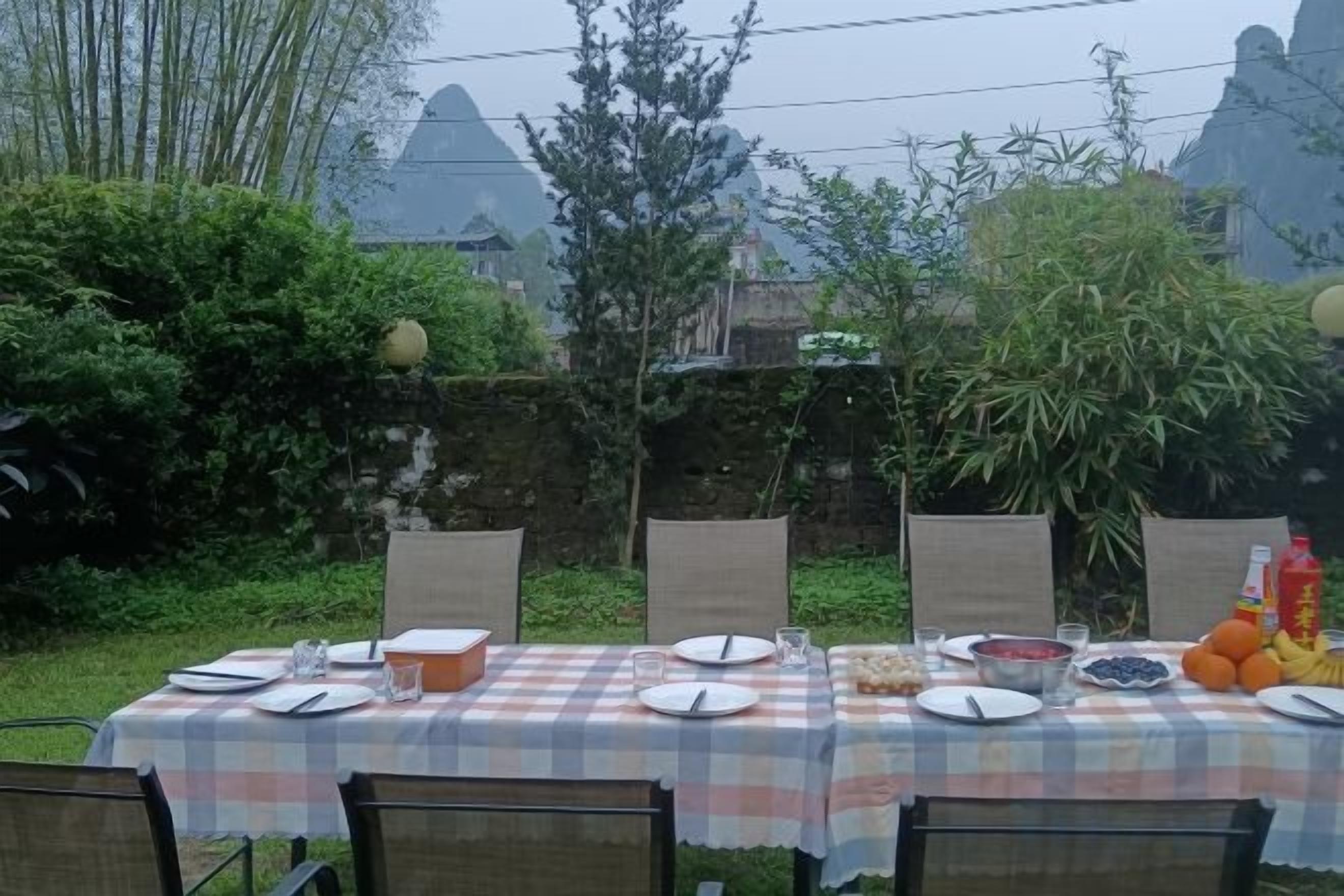 Uma área de refeições ao ar livre com uma mesa posta para quatro pessoas, rodeada por cadeiras, com um muro de pedra e vegetação ao fundo.