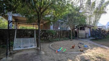 Patio trasero cercado con zona de juegos de arena, un cobertizo pequeño y un árbol que da sombra.