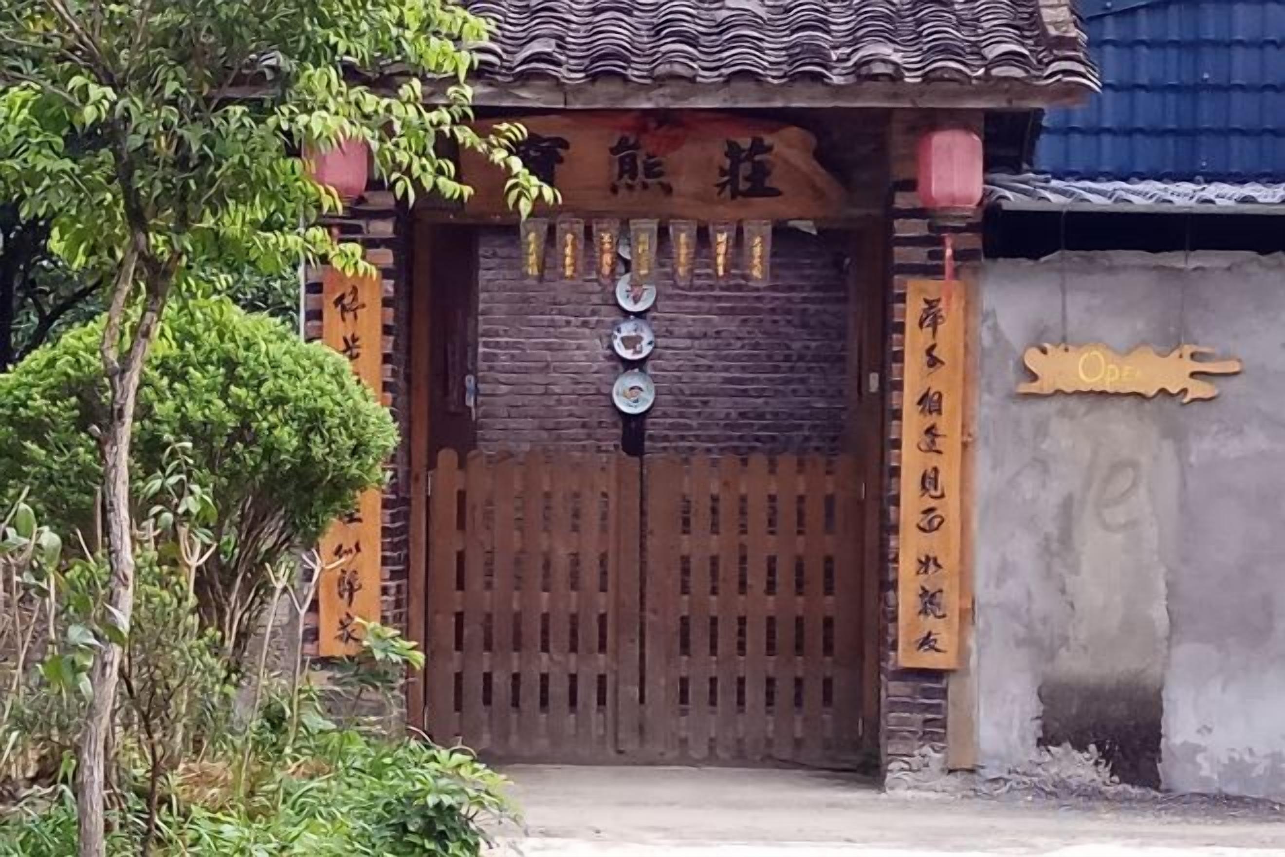 A traditional gate with a wooden design, a sign with Chinese characters, and a small tree to the left.