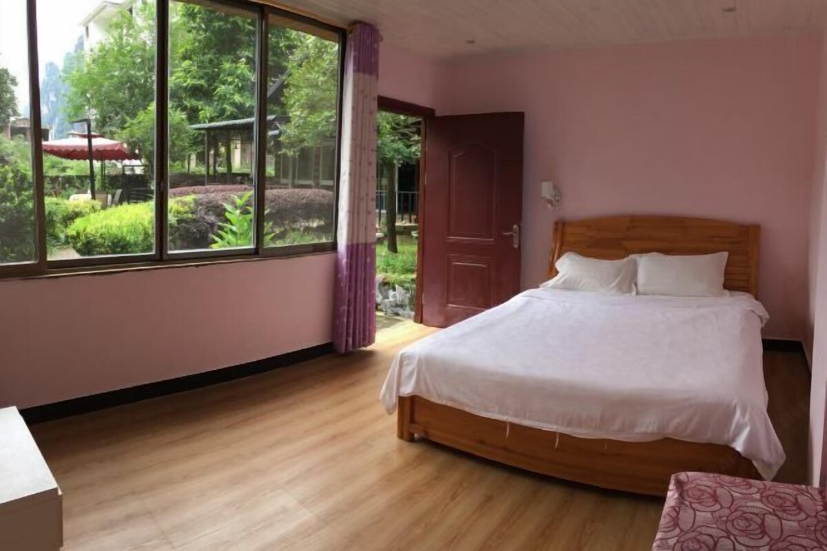 A bedroom with a wooden bed, a large window with a view of greenery, and a door leading outside.