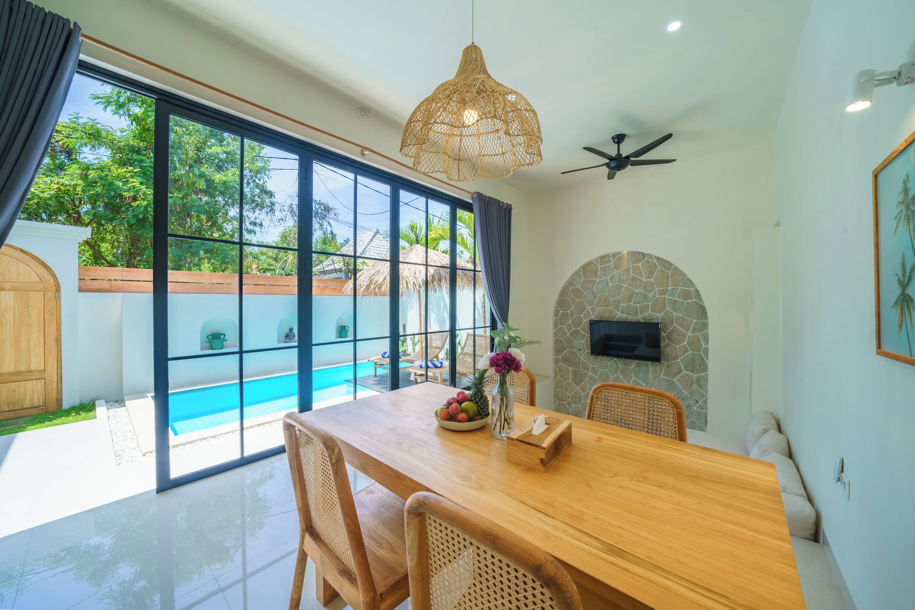 A dining area with a wooden table, wicker chairs, a fruit bowl, and a vase with flowers.