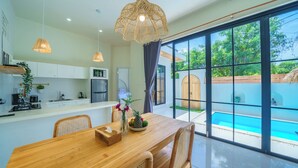 Una cocina moderna con armarios blancos, una mesa de comedor de madera, sillas de mimbre y un amplio ventanal con vistas a una piscina.