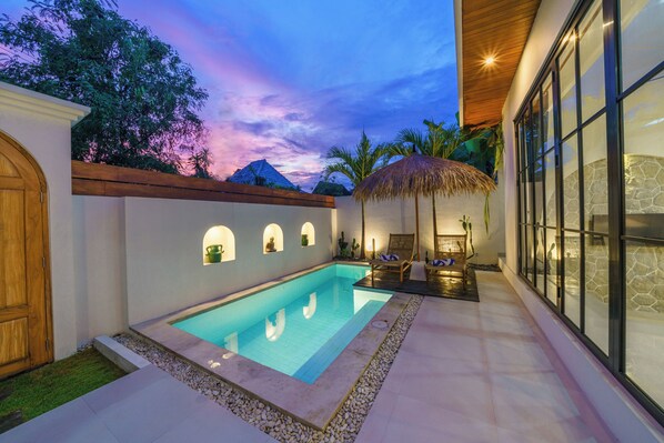A swimming pool with a stone border, a wooden door, and a thatched umbrella. - Villa Selina (Canggu)