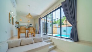 Una sala de estar moderna con sofá, mesa de comedor y sillas. Cuenta con un ventanal con cortinas, una escalera y vistas a una piscina exterior.