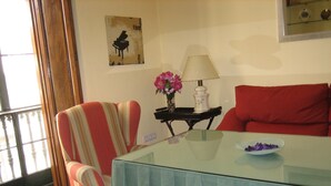 Una sala de estar con una mesa de vidrio, sillones a rayas, un sofá rojo, un cartel de un piano, un jarrón con flores y una lámpara.