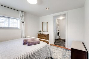 A bedroom with a bed, a dresser, a mirror, a window with curtains, and a laundry area with a washing machine.