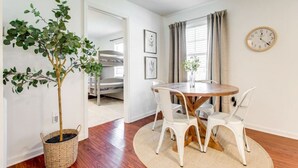 A room with a round wooden table, white chairs, a potted plant, a clock on the wall, and a bunk bed in the background.