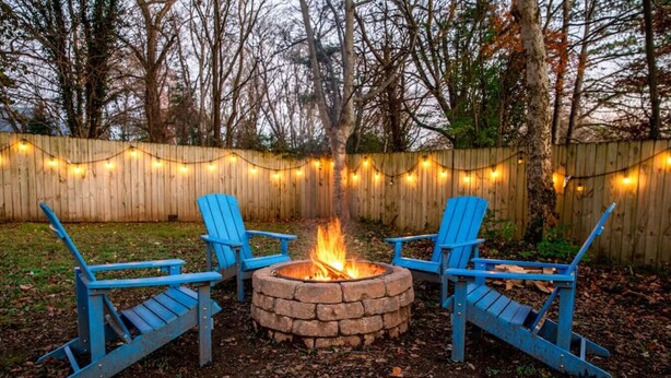 A backyard with a fire pit, three blue Adirondack chairs, and a wooden fence.