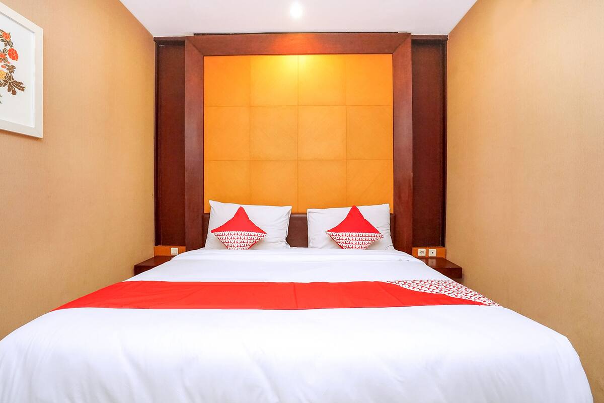 A hotel room with a large bed, two pillows, a red and white bedspread, and a wooden headboard.