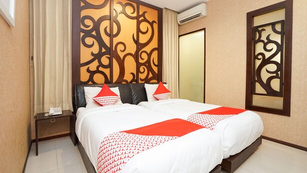 A hotel room with a large bed, a nightstand with a phone, a decorative wall panel, and an air conditioning unit. - Istana Permata Ngagel (Surabaya)