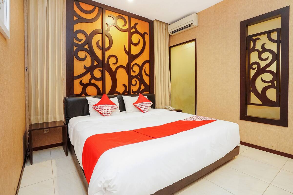 A hotel room with a large bed, a wooden headboard with intricate carvings, a red and white bedspread, and a small bedside table.