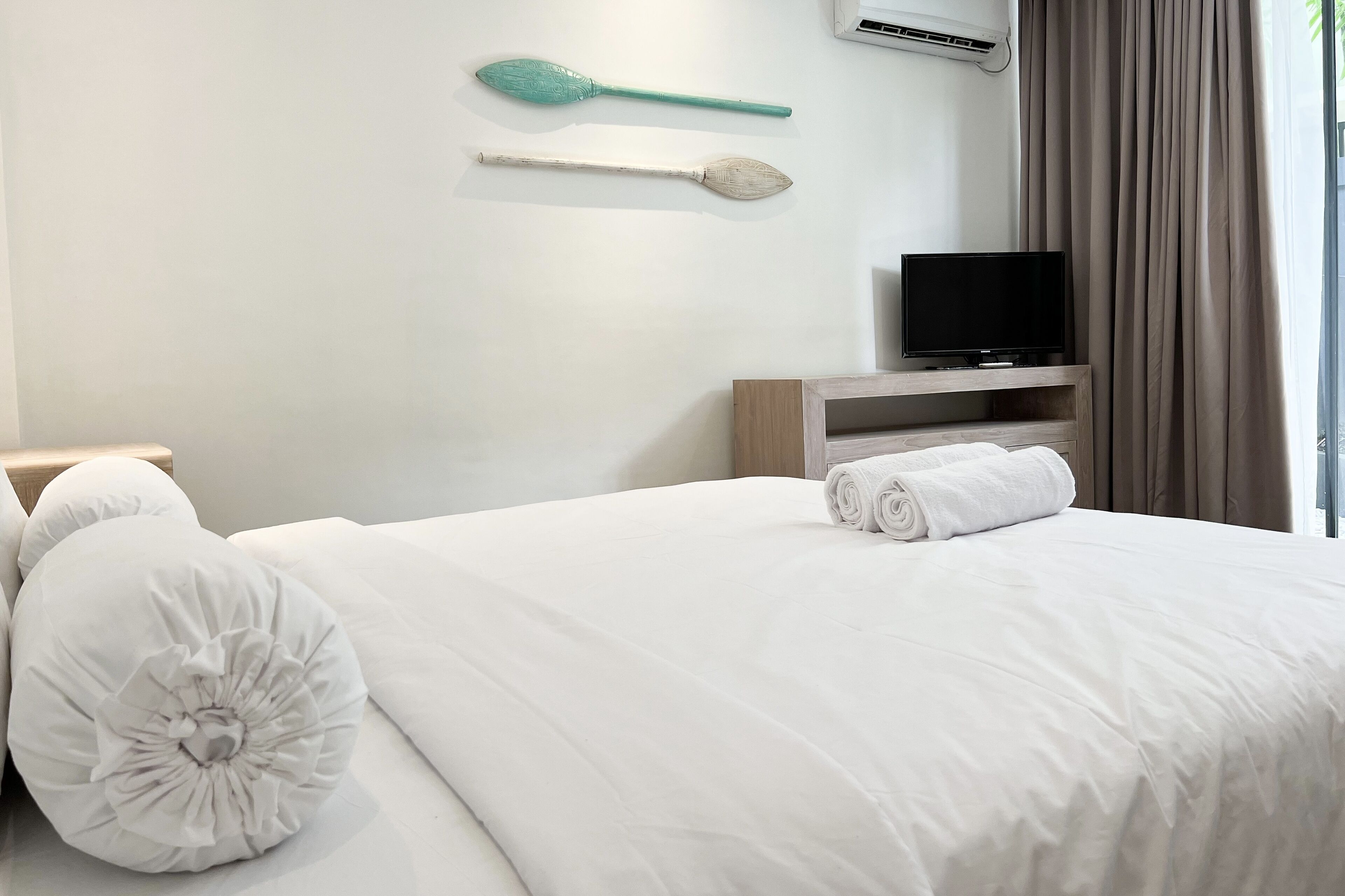 A neatly made bed with white bedding and pillows, a television on a wooden stand, and a wall-mounted air conditioner.