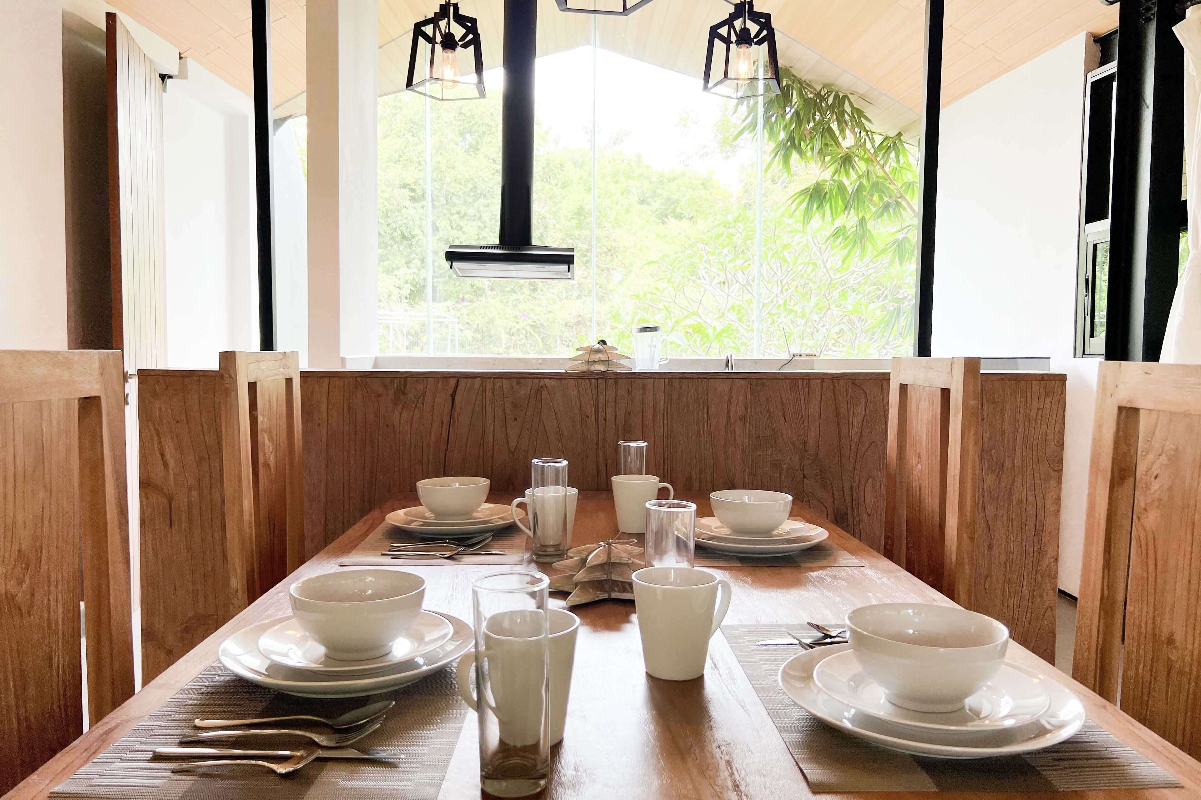 A dining table set with white bowls, glasses, and plates, with a view of greenery outside.