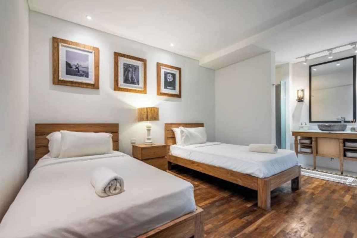 A hotel room with two beds, a wooden floor, a desk with a mirror, and framed pictures on the wall.
