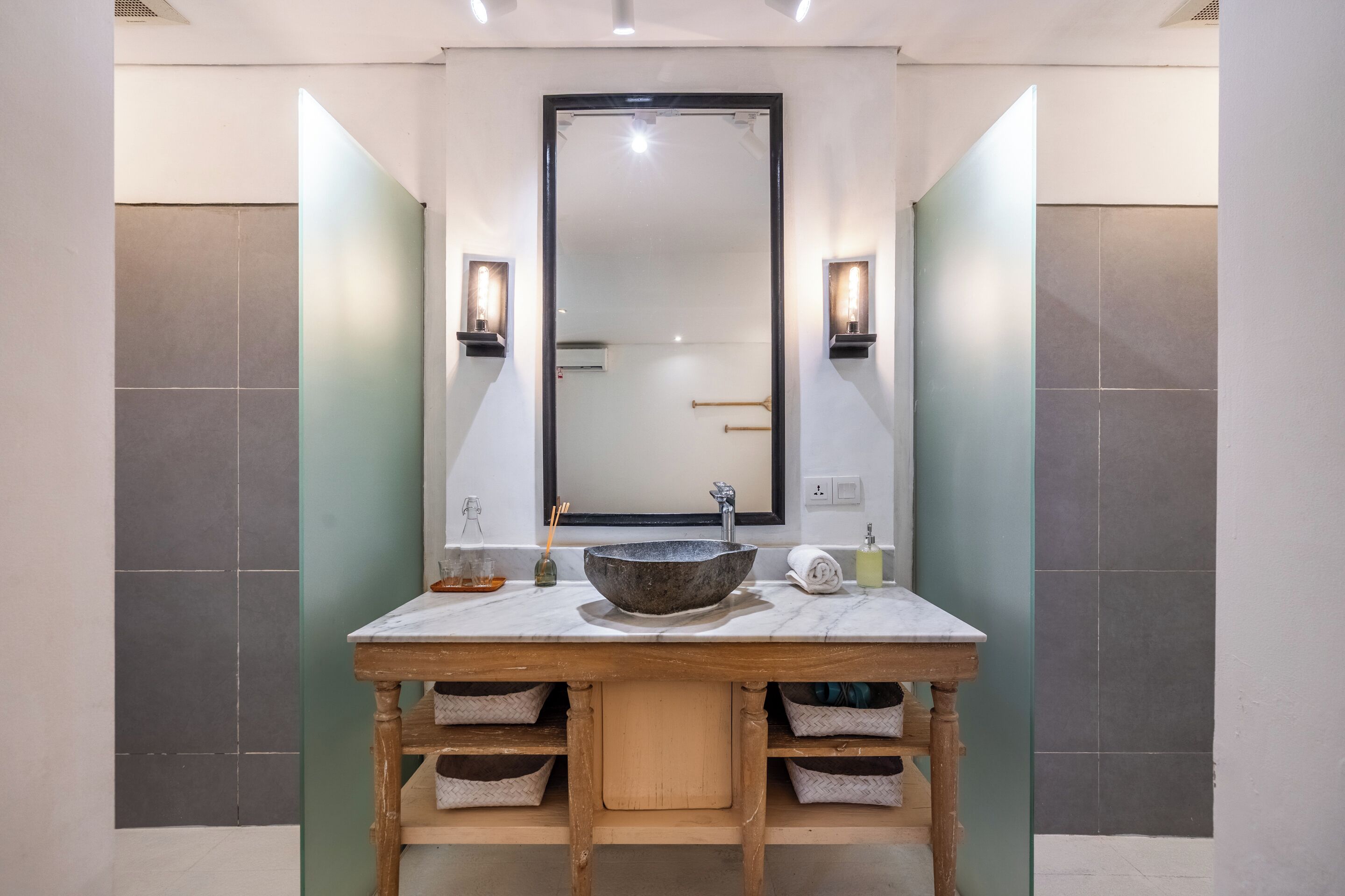 Un baño con un espejo grande, un lavabo de piedra, repisas de madera e iluminación en la pared.