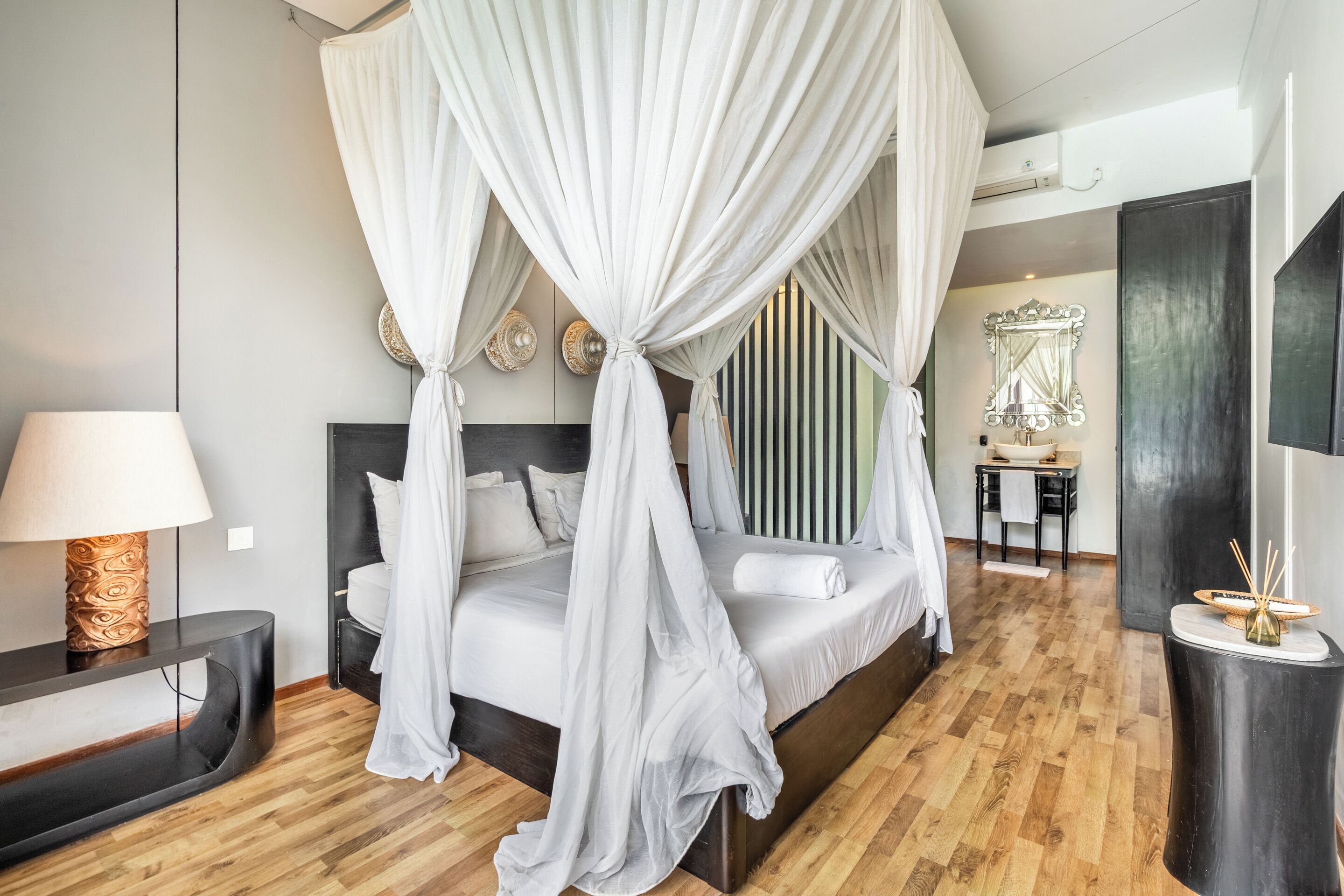 A modern bedroom with a four-poster bed, a bedside table with a lamp, a wooden floor, and a large mirror above the bed.