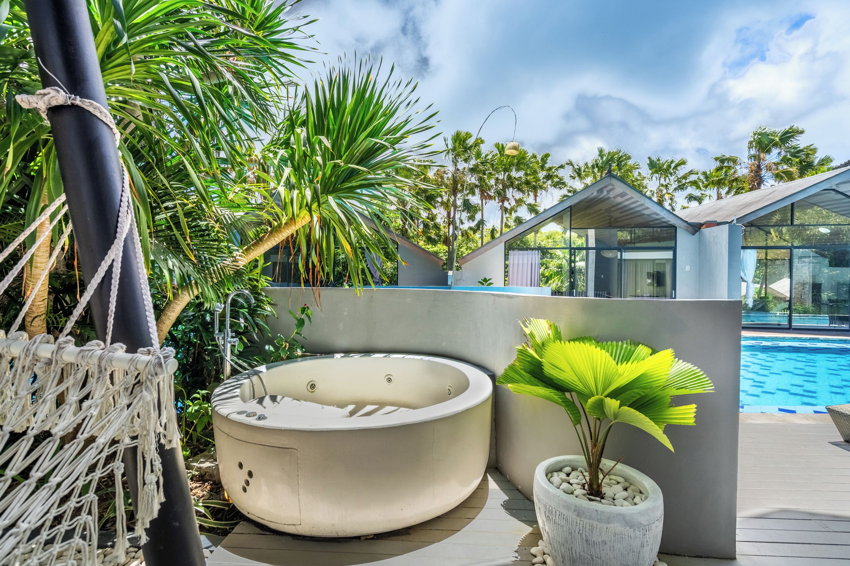 A hot tub, a macrame chair, a potted plant, and a poolside area with palm trees.
