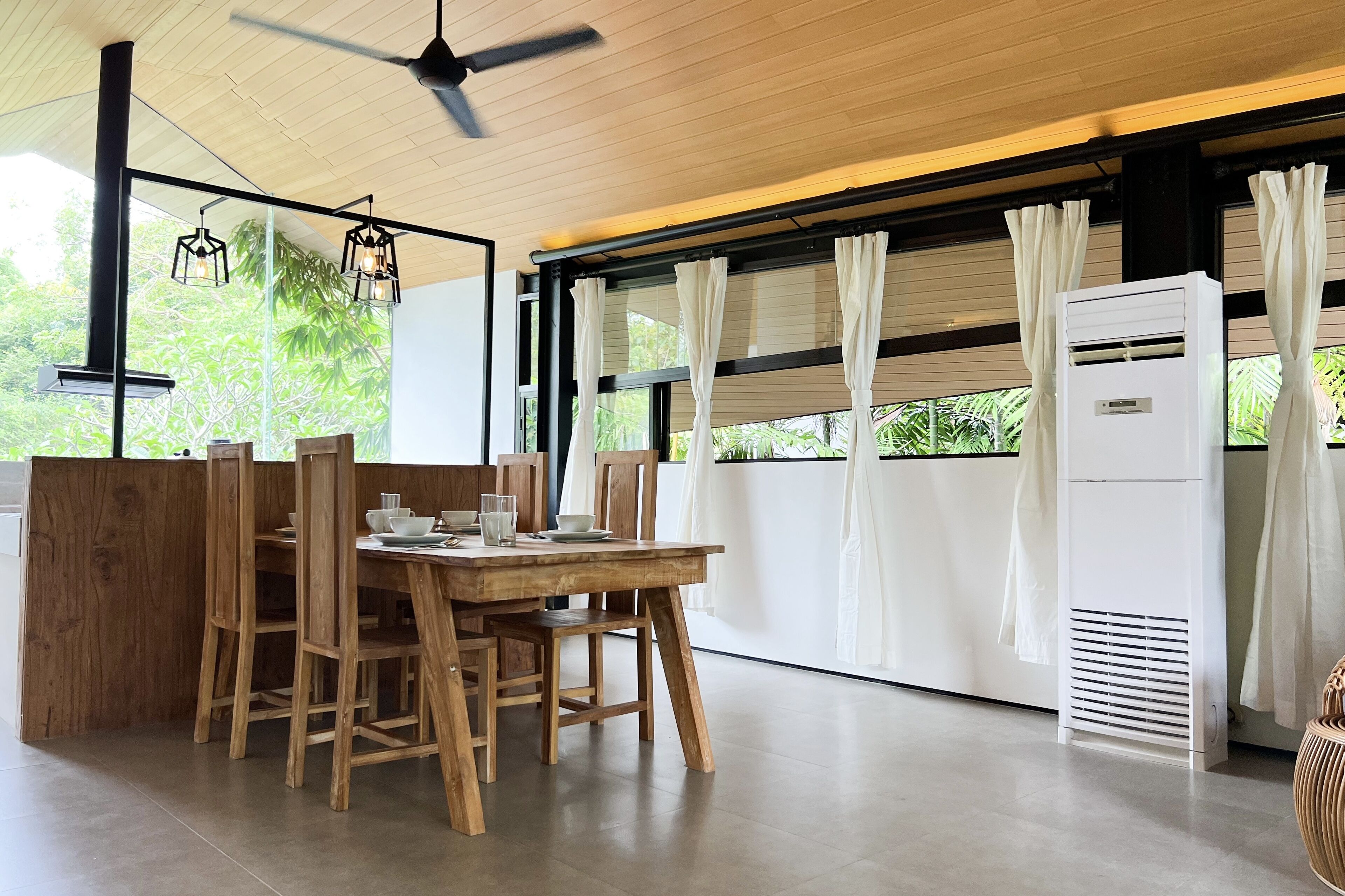 A dining area with a wooden table and chairs, a ceiling fan, and a view of greenery through large windows.